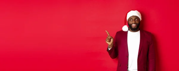 stock image Christmas, party and holidays concept. Cheerful Black man smiling, pointing fingers left and looking at logo, standing over red background.