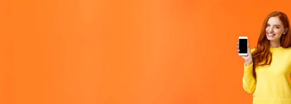 红头发的高加索女孩在智能手机上展示应用程序 如手持手机和信用卡 使用移动银行应用程序 开心地微笑相机 在橙色背景上做广告 — 图库照片
