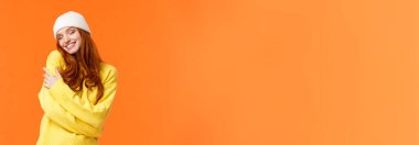 Şefkat, kış tatili konsepti. Çekici, kızıl saçlı, yumuşak kazaklı, beyaz bereli, kapalı gözlü ve kendi vücudunu kucaklayan, kendine sarılan, neşeli, turuncu arka planda gülümseyen bir kadın..