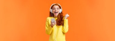 Yaşasın evet yeni şarkı. Çekici, neşeli ve heyecanlı kızıl saçlı kadın. Neşeli ve olumlu duygularla yumruk tokuşturur, kulaklık takar, akıllı telefon tutar, mutlu bir şekilde gülümser kamera, turuncu arka plan.