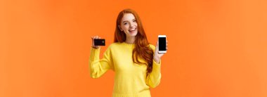 Alışveriş, online ve finans konsepti. Neşeli genç kızıl saçlı kadın banka hesabı açtı, elinde akıllı telefon ve kredi kartı var, gülümsüyor, online alışveriş başvurusu öneriyor..