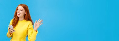 Sevimli ve arkadaş canlısı, kızıl saçlı, akıllı telefon tutan, mesaj atan ve birine dikkat çekmesi için el sallayan, merhaba diyen, gülümseyen, mavi arka planda duran bir kadın. Boşluğu kopyala