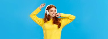 Kaygısız, sarı kazaklı, neşeli ve rahat dans eden, elleri havada, gözleri kapalı, mutlu gülümseyen kulaklıklı, müzik kulaklıklı, mavi arka planda duran çekici bir kadın..
