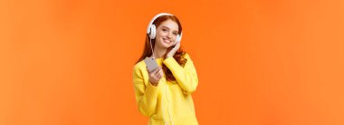 Belden yukarı, neşeli, kızıl saçlı güzel bir kadın. Kulaklıkla müzik dinle, başını eğ ve gülümse. Akıllı telefon tut, sakin bir akşam için şarkı seç, turuncu arka plan..