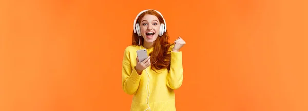 ホーリーはい新曲です 楽しさと肯定的な感情で魅力的な陽気で興奮した赤毛の女性拳ポンプ ヘッドフォンを身に着けています スマートフォンを保持します 幸せな笑顔カメラ オレンジの背景 — ストック写真