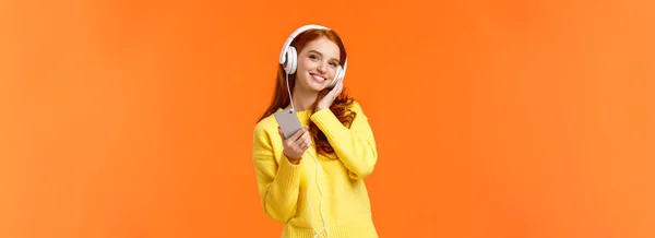 ウエストアップの肖像画明るい赤髪の若い女性は ヘッドフォンで音楽を聞き 頭を傾け 満足笑みを浮かべて スマートフォンを保持し 柔らかい穏やかな夜のために歌を選ぶ オレンジの背景 — ストック写真