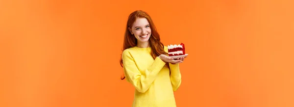 Cute friendly redhead woman holding tasty home-made piece cake and smiling, share bite, treat friends, grinning as celebrating birthday or like eating sweet delicious desserts, orange background.
