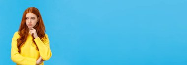 Üzgün, çekingen ve yalnız kızıl saçlı kadın da hayal kırıklığına uğramış ve bunalımda, üzgün ve kıskanç bakıyor, kaşlarını çatıyor, dudağına dokunuyor, kötü hissediyor, mavi arka planda endişeli duruyor..