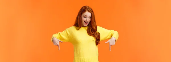 驚きと興奮 指を指して黄色のセーターで面白いかわいい赤毛の女性 下を見て笑顔 チェックアウト製品プロモーション 冬の休日のイベント オレンジの背景 — ストック写真