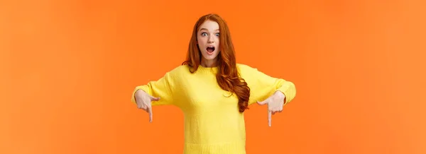 早く見てください 驚くほどの魅力的なかわいい生姜の女の子 黄色のセーターの赤毛の学生は 驚きと不思議から口を開けて 指を指して 見て感銘を受け オレンジの背景 — ストック写真