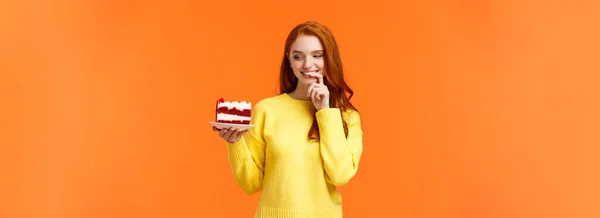 陽気で満足し 幸せな赤毛の女性がカンニングしている日おいしい食べ物を食べて おいしい作品ケーキを保持し 唇をかむと笑顔 誘惑に抵抗することはできません オレンジの背景をかむ — ストック写真