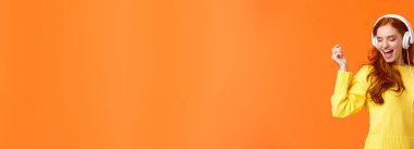 Teknoloji, duygular ve alet konsepti. Neşeli, güzel, kızıl saçlı, elleri havada, gözleri kapalı, neşeyle gülümseyen, kulaklıkla harika müzik seslerinin keyfini çıkaran, akıllı telefon tutan bir kadın..