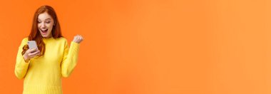 Yeni resimde binlerce beğeni almış bir kız. Neşeli ve zafer kazanan güzel kızıl saçlı kadın yumruk pompası evet de, tezahürat yap ya da kutlama yap, akıllı telefon ekranı, turuncu arka plan.