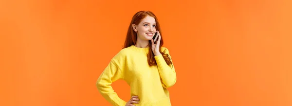 挺拔的肖像漂亮红头发的女人打电话给朋友 聊着天 在假日购物时确认网上订购单 站在旁边无忧无虑地拿着智能手机 橙色背景 — 图库照片