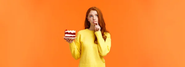 忧心忡忡 忧心忡忡的红头发少女 照顾着自己的身体 计算卡路里 想吃美味的蛋糕 拿着甜点 抬起头去想心事缠身的橙色墙壁 — 图库照片