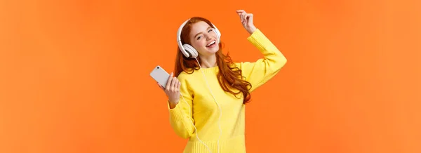 ケアフリー幸せな赤毛十代の女の子は クリスマスプレゼントのための新しいヘッドフォンを受信します 好きな音楽を聞きながらダンスリフトの手を上げます スマートフォンを保持します 置く素晴らしいトラック オレンジの背景 — ストック写真