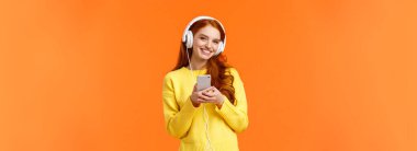 Teknoloji, alışveriş ve insan konsepti. Beyaz kulaklık takan, elinde akıllı telefon tutan, kaygısız gülümseyen, güzel müziğin tadını çıkaran turuncu arka planda duran modern, sevimli hippi kız..
