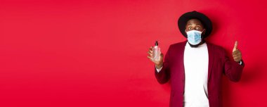 Covid-19, karantina ve tatil konsepti. Etkilenmiş siyah adam parti kıyafeti ve tıbbi maske takıyor, baş parmağını kaldırıyor ve el dezenfektanını gösteriyor, kırmızı arka planda duruyor..