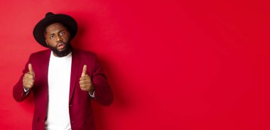 Etkileyici siyahi adam iyi iş çıkarıyor, başparmaklarını kaldırıyor, bir şeyleri övüyor, kırmızı arka planda duruyor. Boşluğu kopyala