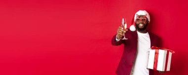 Noel zamanı. Parti gözlüğü ve Noel Baba şapkası takan yakışıklı Afro-Amerikan erkeği elinde yeni yıl hediyesi ve bir bardak şampanya tutuyor, mutlu tatiller diliyor, kırmızı arka plan.