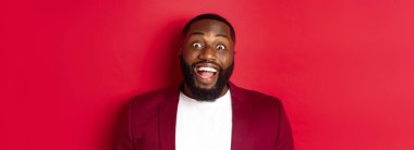 Afrika kökenli, sakallı, kameraya hayran kalmış, gülümseyen ve heyecanlı görünen, ceketle kırmızı arka planda duran Amerikalı adamın yakın çekimi..