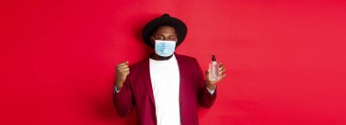 Covid-19, karantina ve tatil konsepti. Neşeli siyah adam kazanmaktan, hedefe ulaşmaktan, antiseptik göstermekten, tıbbi maske takmaktan, kırmızı arka planda durmaktan memnun..