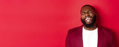 Mutlu ve kaygısız siyahi adamın yakın plan çekimi. Eğleniyor, gülüyor ve gülümsüyor. Ceketin içinde kırmızı arka plana karşı duruyor..