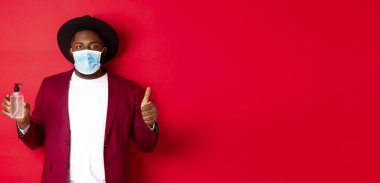 Covid-19, karantina ve tatil konsepti. Tıbbi maskeli, el dezenfektanı ve baş parmağını kaldırmış, antiseptik kullanmayı öneren Afrikalı genç bir Amerikalı..