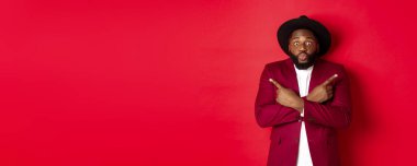 Noel alışverişi ve insanların konsepti. Kararsız siyah adam iki değişken arasında seçim yapıyor, parmakları yana doğru gösteriyor ve kafası karışmış kameraya bakıyor, kırmızı arka plan.