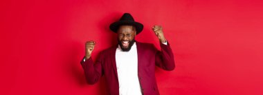 Başarılı takım elbiseli siyahi adam evet diyor, kazanmanın veya hedefe ulaşmanın sevinciyle el kaldırıyor ve sevinç çığlıkları atıyor, kırmızı arka plan.