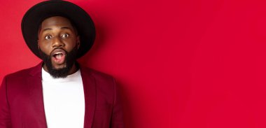 Etkilenmiş, sakallı, kameraya bakan, çenesini indiren ve kırmızı arka planda duran siyahi adamın yakın çekimi..