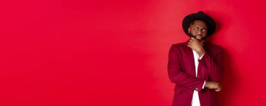 Moda ve parti konsepti. Şüpheci ve şüpheci Afro-Amerikan adam düşünüyor, sol üst köşede hoşnutsuz görünüyor, şüpheleri var, kırmızı arka plan.