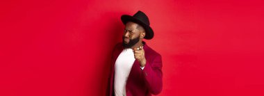 Şüpheli siyah adam kamerayı işaret ediyor ve gözlerini kısıyor, seni suçluyor, uyarı veriyor, kırmızı arka planda duruyor..