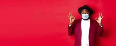 Covid 19 ve moda konsepti. Şapkalı ve ceketli şık Afro-Amerikan adam, yüz maskesi takıyor ve kırmızı arka planda duruyor..
