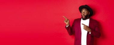 Kış tatili ve alışveriş konsepti. Mutlu siyah adam solu işaret ediyor ve gülümsüyor, kırmızı arka planda yeni yıl tanıtım teklifini gösteriyor..