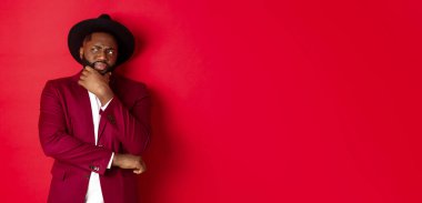 Moda ve parti konsepti. Şüpheci ve şüpheci Afro-Amerikan adam düşünüyor, sol üst köşede hoşnutsuz görünüyor, şüpheleri var, kırmızı arka plan.