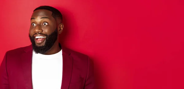 stock image Close-up of excited african american guy looking left, smiling with amazement and joy, standing in blazer against red background.