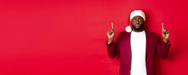 stock image Christmas, party and holidays concept. Upset african-american guy in santa hat complaining, pointing fingers up and frowning disappointed, standing against red background.