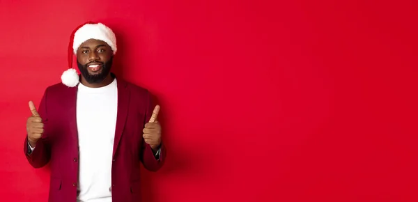 stock image Christmas and party concept. Handsome Black man celebrating winter holidays, wearing santa hat and showing thumb up, recommending new year offer, red background.