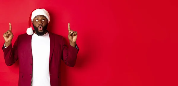 stock image Christmas, party and holidays concept. Impressed Black man in santa hat pointing fingers up, saying wow and staring at camera, checking out promo offer, red background.