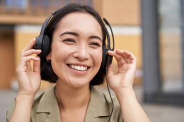 Kulaklıklı güzel Asyalı bir kadının portresi, şehir merkezinde müzik dinliyor, mutlu bir şekilde gülümsüyor..