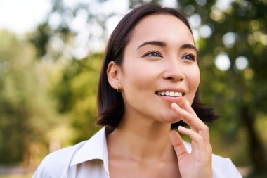 Genç Asyalı kadın portresi, gülüyor ve gülümsüyor, parkta yürürken mutlu görünüyor, yeşil ağaçların yanında poz veriyor..