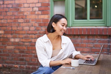 Şık bir genç iş kadını, laptoplu Asyalı bir kız, bir fincan kahveyle açık kafede oturuyor ve çalışıyor, bilgisayar kullanıyor..