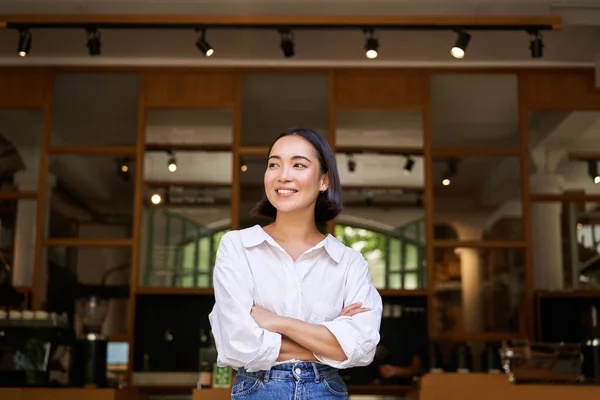 stock image Portrait of confident young asian woman, business owner, cross arms on chest, looking pleased, standing in front of cafe.