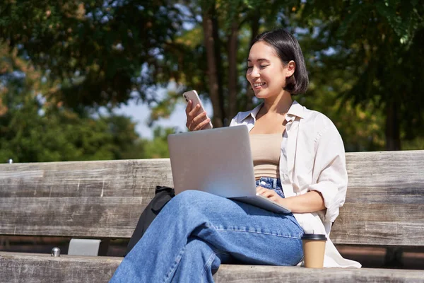 stock image Excited young asian girl, looking at her smartphone, while sitting with laptop outdoors in sunny park.