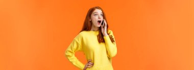 Şaşırmış, ürkmüş güzel kızıl saçlı kadın telefonda konuşurken inanılmaz haberler duyuyor, çenesini indiriyor sola bakıyor ve konuşma sırasında akıllı telefonu kulağına yakın tutuyor, turuncu arka planda duruyor..