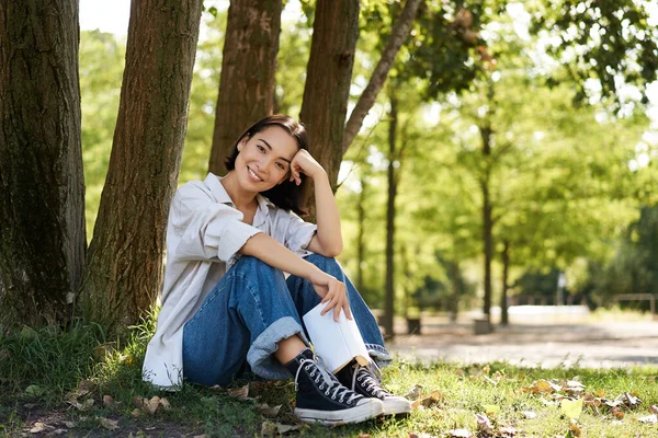 stock image Woman sitting in park with her favourite book, leaning on tree under shade on sunny day, enjoying nature and calm relaxing atmosphere.