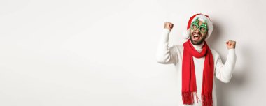 Noel, yeni yıl ve kutlama konsepti. Parti gözlüklü ve Noel Baba şapkalı mutlu erkek manken. Yumruk sallıyor ve zafer kazanıyor. Beyaz arka planda duruyor..