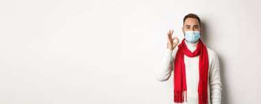 Covid-19, tecrit ve karantina konsepti. Yüz maskesi takmış tatmin olmuş yakışıklı adam işaret veriyor, onaylıyor ya da katılıyor, beyaz arka plan gibi..