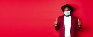Covid 19 ve moda konsepti. Şapkalı ve ceketli şık Afro-Amerikan adam, yüz maskesi takıyor ve başparmağını kaldırıyor, kırmızı arka planda duruyor..